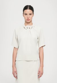 Blusa bianca a maniche corte realizzata in tessuto morbido, con scollo plissettato decorato da tre bottoni metallici e una vestibilità comoda.