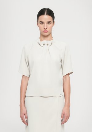 Blusa bianca a maniche corte realizzata in tessuto morbido, con scollo plissettato decorato da tre bottoni metallici e una vestibilità comoda.