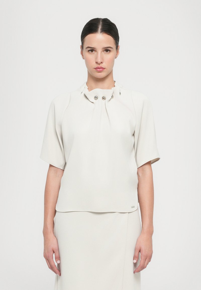 Blusa bianca a maniche corte realizzata in tessuto morbido, con scollo plissettato decorato da tre bottoni metallici e una vestibilità comoda.