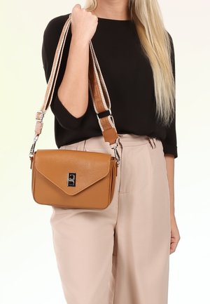 Frau trägt schwarzen Top und beige Hose und hält eine hellbraune Leder-Umhängetasche mit gestreiftem, verstellbarem Schulterriemen.