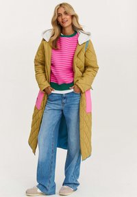 Plyšová žltá prešívaná bunda s ružovými a modrými akcentmi nad ružovým pruhovaným svetrom a modrými džínsami, doplnená béžovými teniskami.