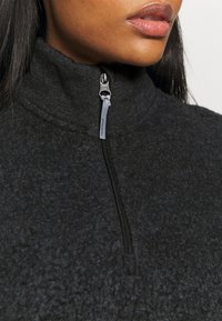 Sort zip-up fleecejakke med en blød tekstur, høj krave og kontrastglidsejr. Hovedstoffet virker tykt og hyggeligt.