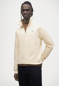 Sweat-shirt zippé couleur crème avec col montant et broderie verte "II", porté sur un haut marron, associé à un jean foncé.