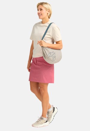 Femme debout portant un t-shirt beige, une jupe rose, des baskets beiges, et tenant un sac à bandoulière gris avec un motif d'empreinte de patte bleue.