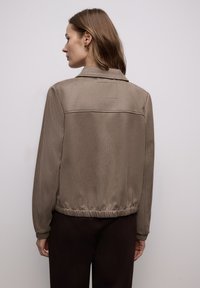 Brun, tekstureret jakke med krave og elastisk taljebånd. Har et enkelt rygdesign med en lille syet mærkedetalje.