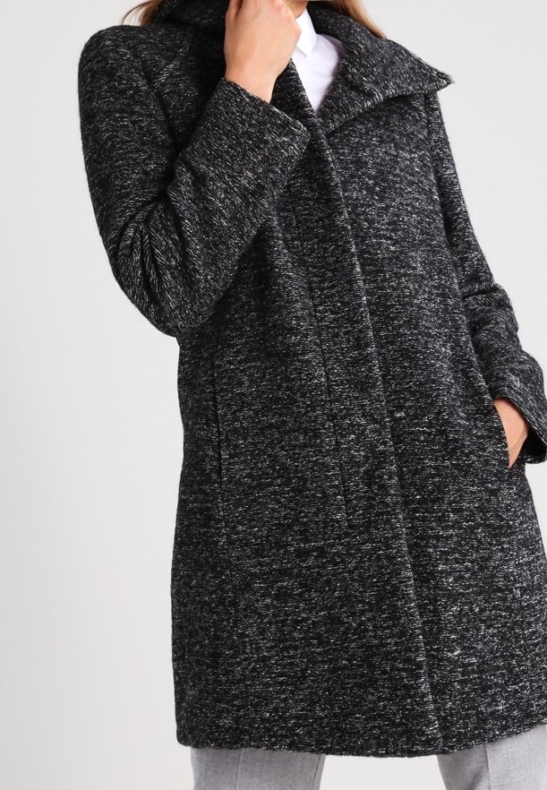 En kvinna håller sin hand nära hakan och bär en stilren svart och grå texturerad kappa som visar sin moderna design mot en ljus bakgrund.