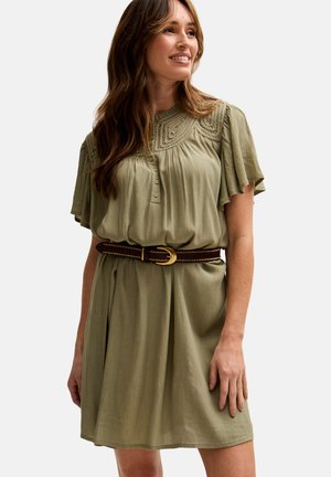 Lächelnde Frau, die ein olivgrünes, kurzärmliges Kleid mit besticktem Passe und einem dunkelbraunen Gürtel mit goldener Schnalle trägt.