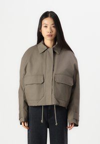 Giacca verde oliva tagliata, realizzata in tessuto resistente, con colletto a punta e due grandi tasche frontali con orlo regolabile.