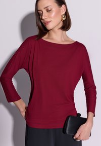 Bordowa bluzka z długim rękawem i dekoltem w łódkę, wykonana z miękkiego materiału, o luźnym kroju, z marszczeniami na ramionach, zestawiona z czarną kopertówką.