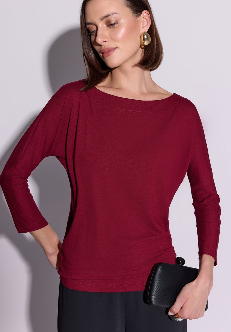 Bordowa bluzka z długim rękawem i dekoltem w łódkę, wykonana z miękkiego materiału, o luźnym kroju, z marszczeniami na ramionach, zestawiona z czarną kopertówką.