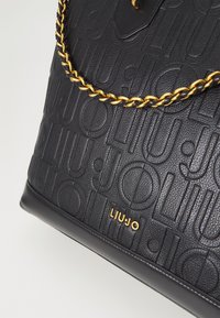 Borsa nera texture con un motivo a logo in rilievo, accentuata da una catena dorata e il nome del marchio "LIUJO" in oro embossato sul davanti.