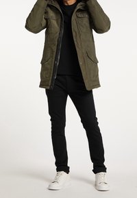 Oliwkowa pikowana parka z zapięciem na zamek i zatrzaski, liczne przednie kieszenie, noszona na czarnej koszuli i czarnych spodniach, zestawiona z białymi tenisówkami.