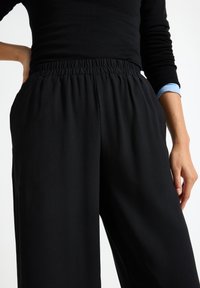Pantalones negros de pierna ancha con cinturilla elástica, con una textura suave de tela y bolsillos laterales. Se vislumbra parcialmente una camiseta negra ajustada.