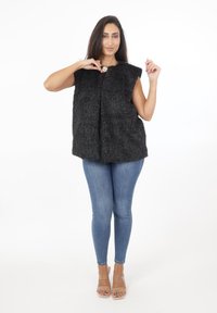 Czarna kamizelka z sztucznego futra z wysokim kołnierzem, otworem z przodu i kieszeniami, w połączeniu z niebieskimi rurkami i przezroczystymi sandałami na obcasie.