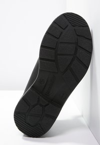 Suela de zapato de goma negra con un patrón de agarre texturizado, que presenta secciones elevadas y detalle de marca. Plana, ancha, con surcos profundos para tracción.