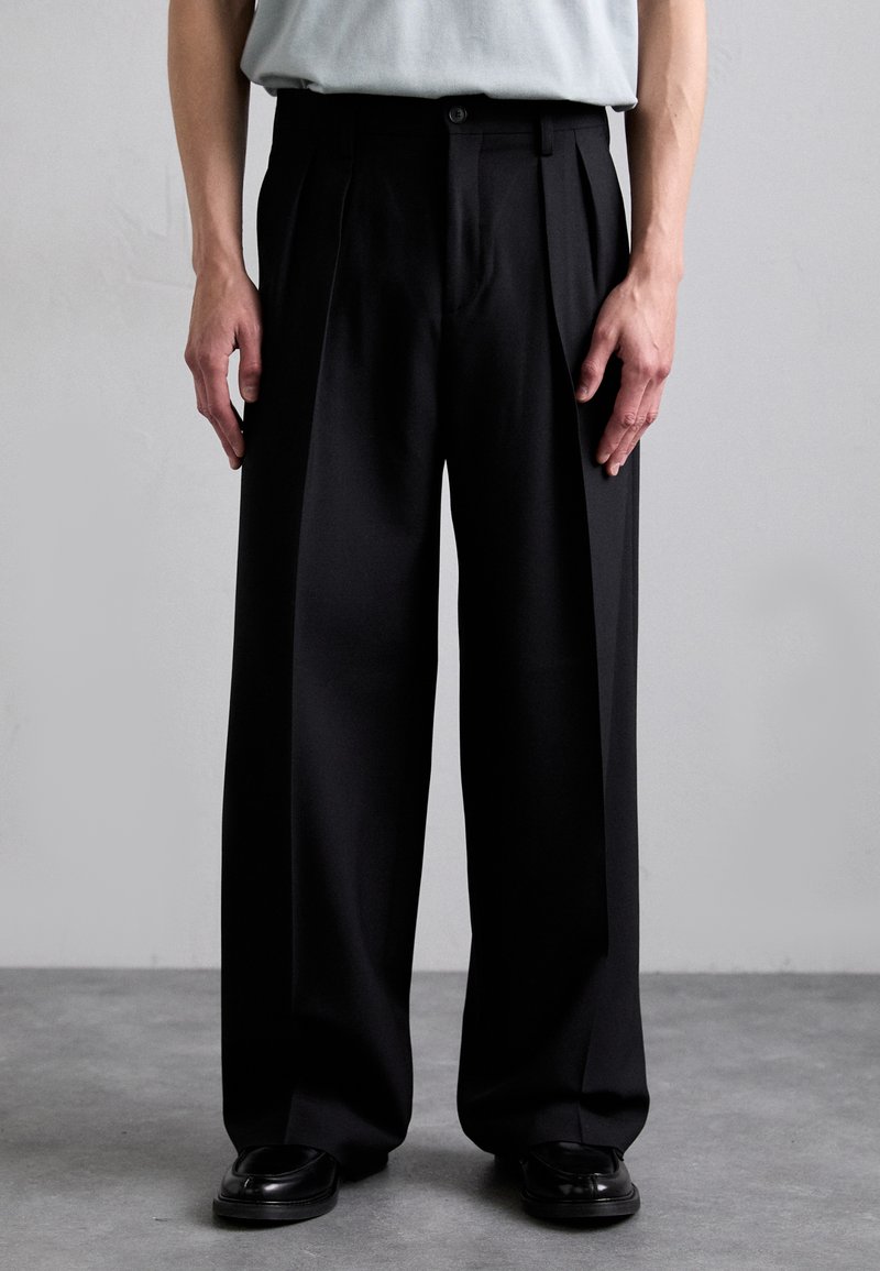 Pantalons plissés noirs en tissu lisse, avec une coupe ajustée, des jambes larges et une fermeture à un bouton à la taille.