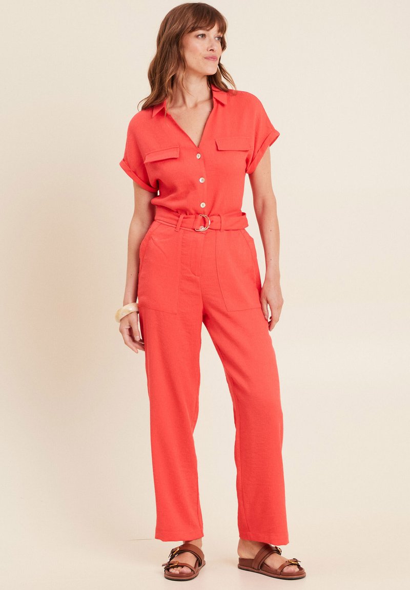 Combinaison corail à manches courtes, deux poches poitrine, taille ceinturée et jambes larges. Texture lisse et fermeture à boutons à l'avant.