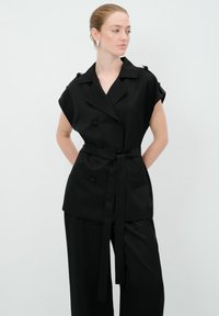 Haut noir à double boutonnage avec des manches courtes, présentant une ceinture à nouer, deux poches avant et des accents de boutons sur les coutures des épaules.