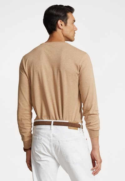 Beige langemouwen shirt gemaakt van zachte stof, gedragen met witte jeans. Het shirt heeft een eenvoudige ontwerp met een relaxte pasvorm en minimale stikseldetails.