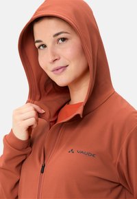 Felpa con zip colore rosso-arancio realizzata in tessuto testurizzato, con cappuccio aderente e un piccolo logo sul lato sinistro. Fodera interna liscia visibile.