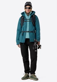 Αδιάβροχο μπουφάν σε χρώμα πετρόλ πάνω από ανοιχτό μπλε ενδιάμεσο στρώμα, σετ με μαύρο παντελόνι και γκρι παπούτσια πεζοπορίας. Συμπληρώνεται με σκουφάκι και γυαλιά ηλίου.