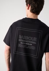 Zwarte katoenen T-shirt met een grote witte grafische tekstprint op de rug, omrand door een rechthoekige contour, met "BARBOUR INTERNATIONAL" en "SOUTH SHIELDS ENGLAND."