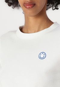 Maglietta a maniche corte bianca con colletto rotondo e un logo ricamato blu di una faccina sorridente con "HGO" sopra, che evidenzia semplicità e comfort.