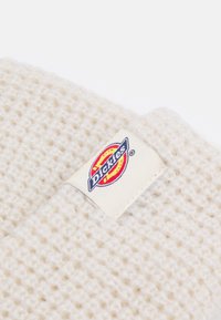 Vävd krämfärgad mössa med ett strukturerat stickat mönster och en sydd Dickies-logotyp i blått, rött och gult på en krämfärgad bakgrund.