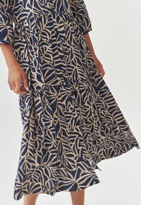 Vestido azul marino y beige con patrones de hojas, cierres de botones, mangas de tres cuartos y un diseño de falda en capas con una textura fluida.