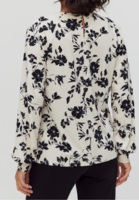 Blumenbluse in Creme mit schwarzen Blumen. Verfügt über lange Ärmel, einen Rundhalsausschnitt und eine Schlüsselhole-Öffnung im Rücken. Weicher Stoff, lockere Passform.