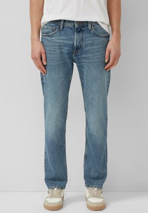 Persoon met rechte blauwe spijkerbroek en witte sneakers, staand tegen een effen lichtgrijze achtergrond.