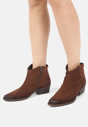 Braune Wildleder-Stiefeletten mit seitlichen Reißverschlüssen, getragen an bloßen Beinen, mit niedrigem Blockabsatz und spitz zulaufender Zehenpartie.