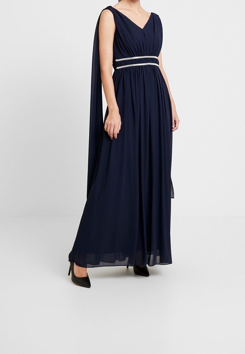 Femme portant une robe longue bleu marine sans manches avec une ceinture argentée et des chaussures à talons noires, debout devant un fond uni.