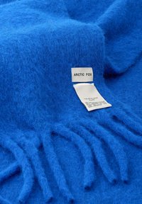 Helderblauwe zachte wol sjaal met franjes aan de uiteinden, voorzien van een wit label met de tekst "ARCTIC FOX" en verzorgingsinstructies.