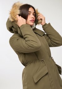 Zielona oliwkowa kurtka puchowa z kapturem wyłożonym sztucznym futrem. Posiada zwężoną talię, dwie przednie kieszenie oraz gładki, teksturowany materiał.