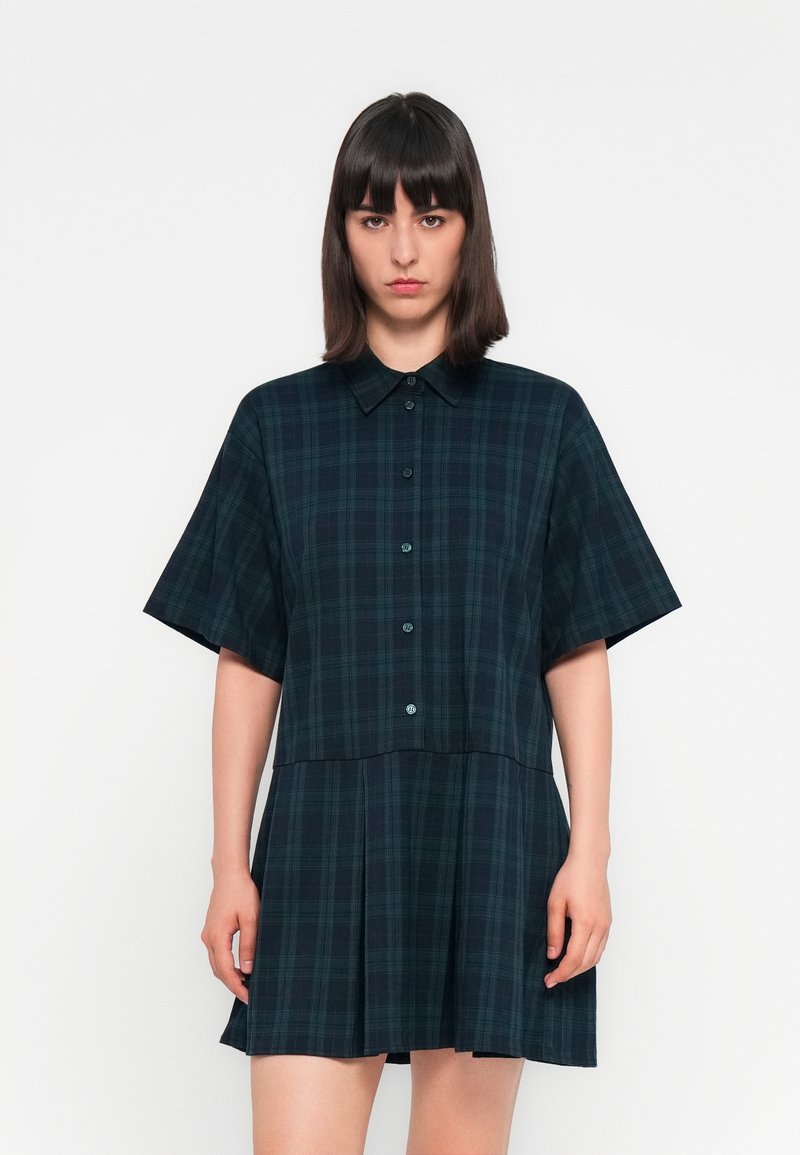 Knielanges Karohemdkleid in Dunkelgrün und Marineblau. Kurze Ärmel, lockere Passform und plissierter Rock. Glattes Material mit klassischem Karomuster.
