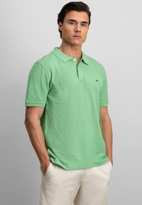 Polo shirt en coton vert avec une patte de boutonnage à deux boutons, manches courtes et un petit logo sur la poitrine gauche. Associé à un pantalon beige clair.