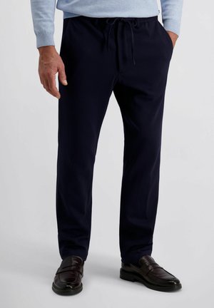 Man in marineblauwe capri-broek, lichtblauwe trui en donkerbruine leren loafers, die met zijn handen in zijn zakken staat.
