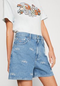 Ljusblå denimshorts med hög midja och broderad "Tommy Jeans"-logga över hela plagget. Har standardfickor och knäppning med knapp.