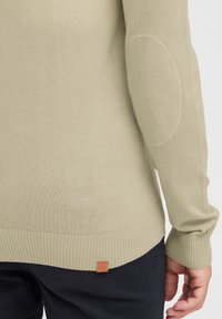 Sweter dzianinowy w jasnym zielonym kolorze o teksturowanym wykończeniu. Posiada ściągacze przy mankietach, u dołu oraz łatki na łokciach, a także małą brązową metkę z logo.