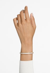 Bracciale in tono oro con un design tessuto e testurizzato e una fila di pietre trasparenti lungo la parte superiore. Indossato su una mano su uno sfondo chiaro.