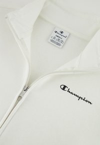 Witte ritsjack met kraag, gemaakt van zachte stof. Bevat een zwart "Champion" logo en een verzorgingslabel aan de binnenkant.