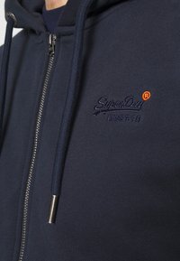 Sudadera con cremallera de color azul marino, con cordones, que presenta un logo bordado de SuperDry y caracteres japoneses en el pecho.