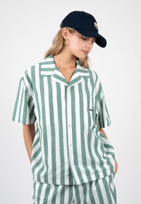 Femme portant une chemise à manches courtes rayée verticale verte et blanche et un pantalon assorti, avec une casquette de baseball bleu marine et des boucles d'oreilles créoles dorées.