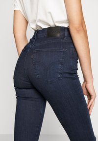 Tmavě modré džíny z denimu s vysokým pasem, obsahující zadní kapsy a koženou nášivku na pase. Hladká textura a přiléhavý střih.