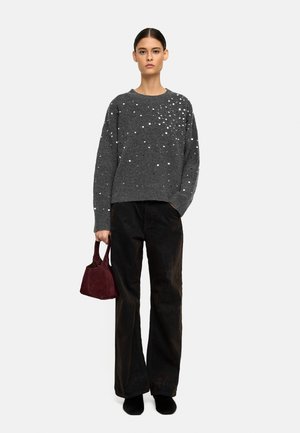 Szary sweter wykonany na drutach z rozrzuconymi białymi zdobieniami, sparowany z czarnymi szerokimi spodniami z korduroju i małą bordową torebką.