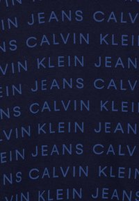 Calvin Klein Jeans upprepas i ett diagonalt mönster med ljusblå text på en mörkblå tygbakgrund.