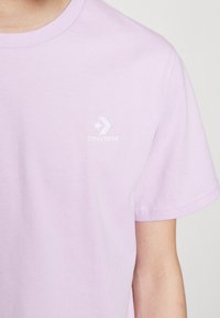 Fialové bavlněné tričko s krátkým rukávem, klasickým kruhovým výstřihem a jemně bílým vyšitým logem Converse na levé straně hrudi. Hladká textura.