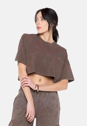 Młoda kobieta z ciemnymi włosami związanymi w kucyk, ubrana w brązowy krótki T-shirt i pasujące spodnie z wysokim stanem, patrzy w lewo.