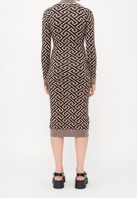 Robe midi ajustée à manches longues avec un motif géométrique en beige et noir représentant un labyrinthe, associée à des chaussures noires à talons larges.